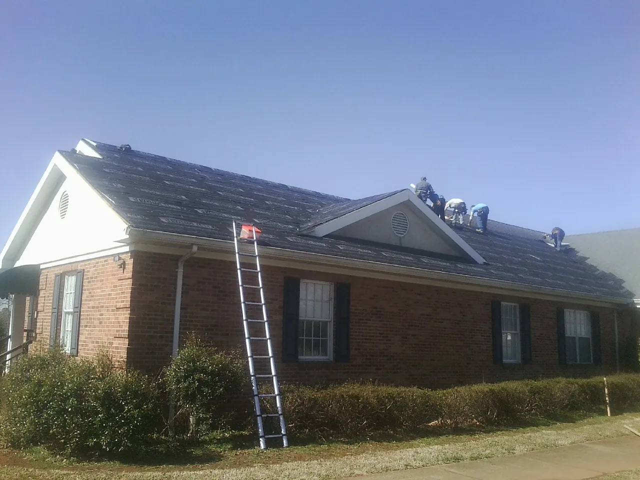 Crew tearing shingles off a brick ranch roof with a ladder set — Emergency Roof Repair work by our crew in Holden